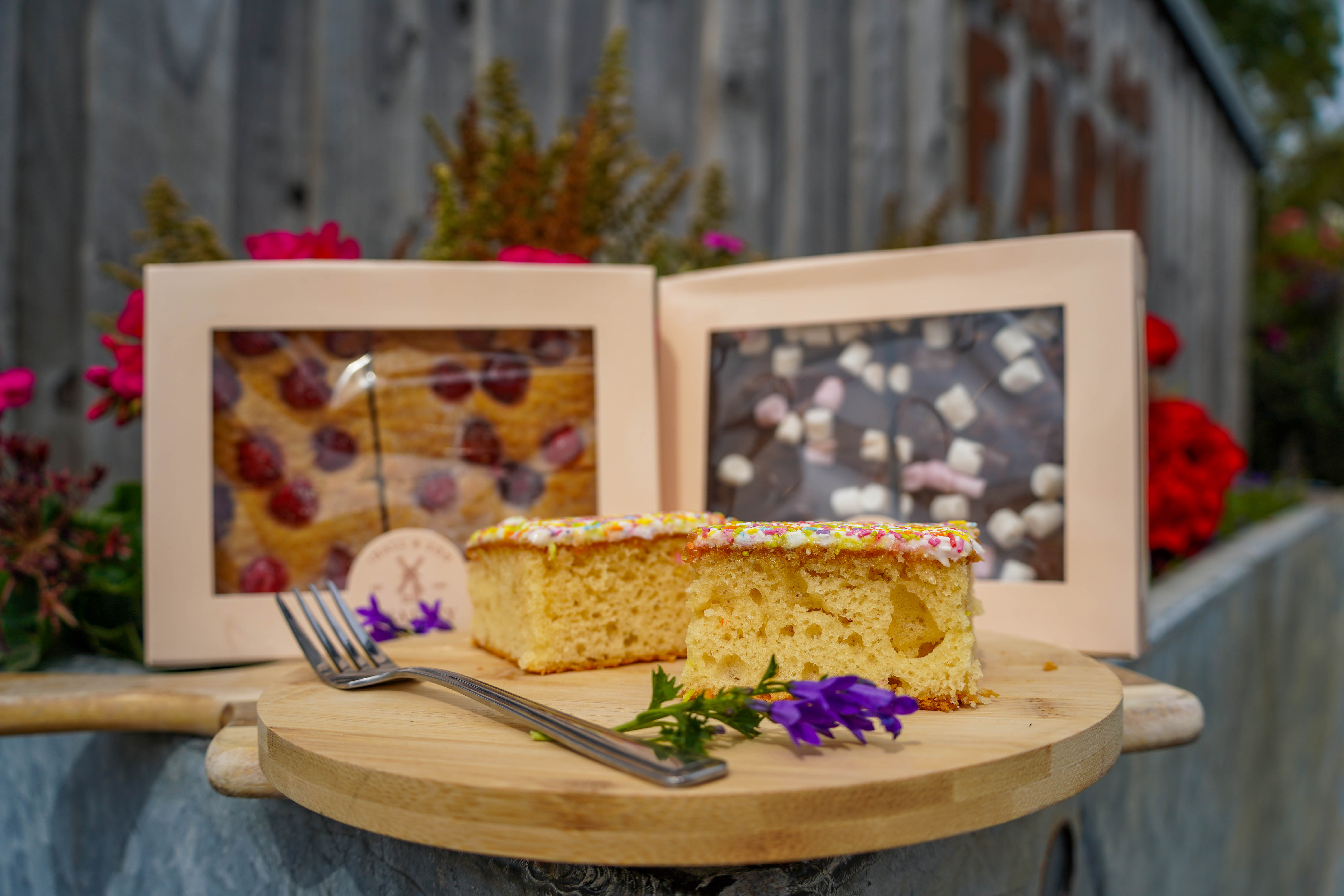 Slices of school cake displayed on wooden board, with boxed cherry shortbread and rocky road behind. 