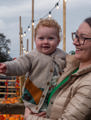 Mum holding child on pumpkin patch with fairy lights in the background as it begins to get dark. 