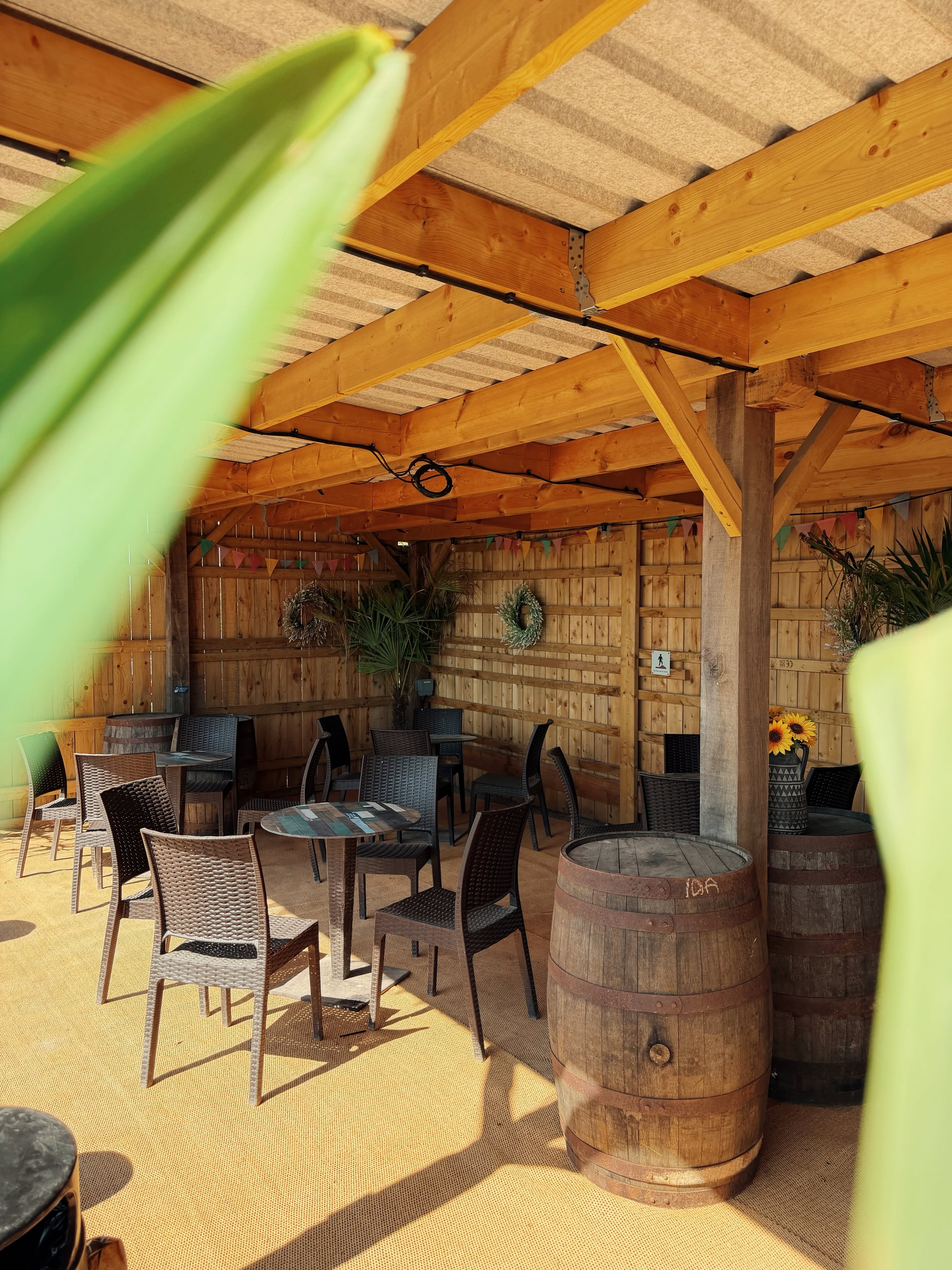 Outside seating area under wooden covering with brown chairs around circular tables and barrels and sunflowers used as decoration. 