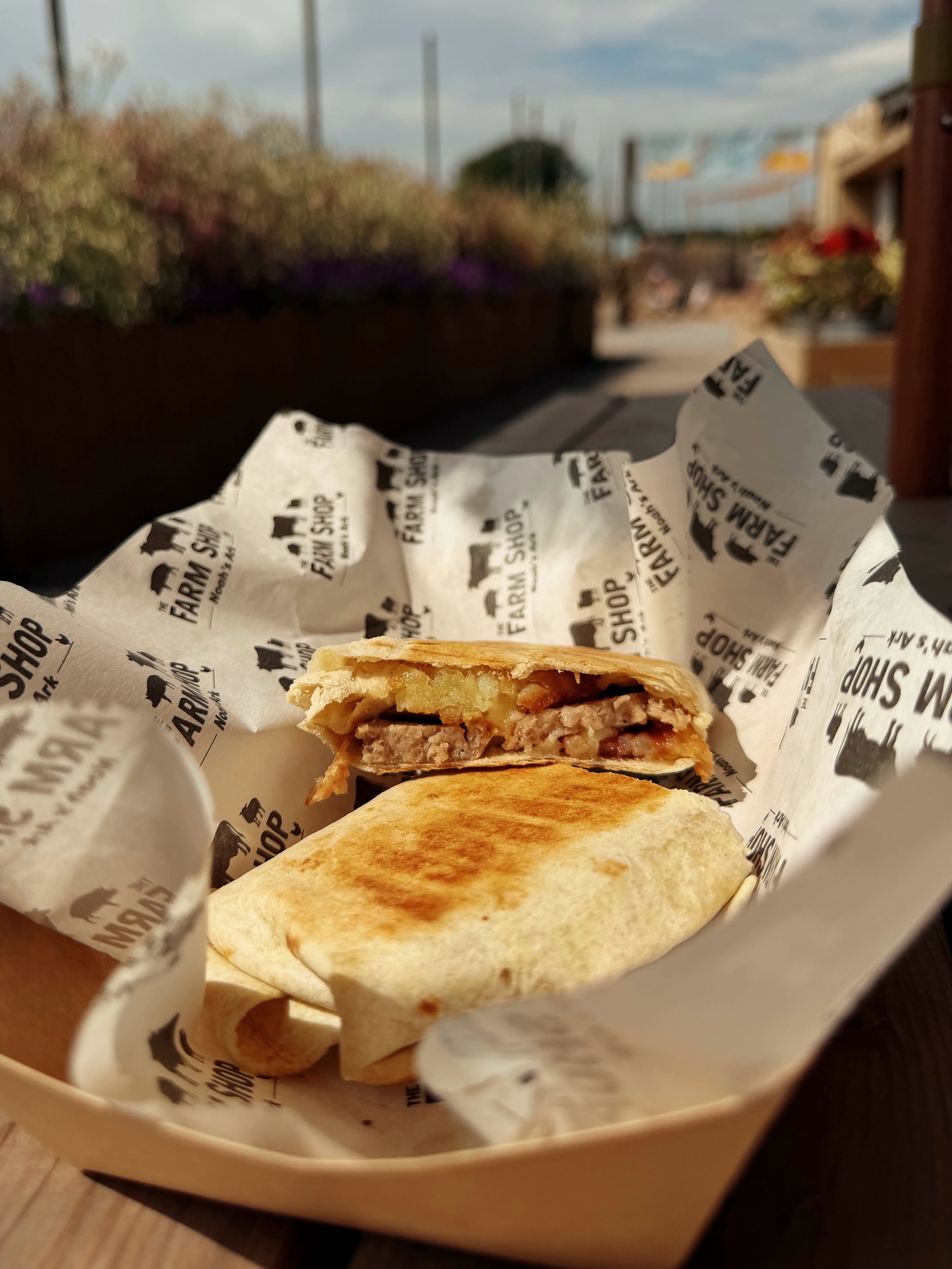 Toasted breakfast burrito, cut in half to show filling and wrapped in Farm Shop branded paper. Photo taken on wooden bench outside the Farm Shop. 